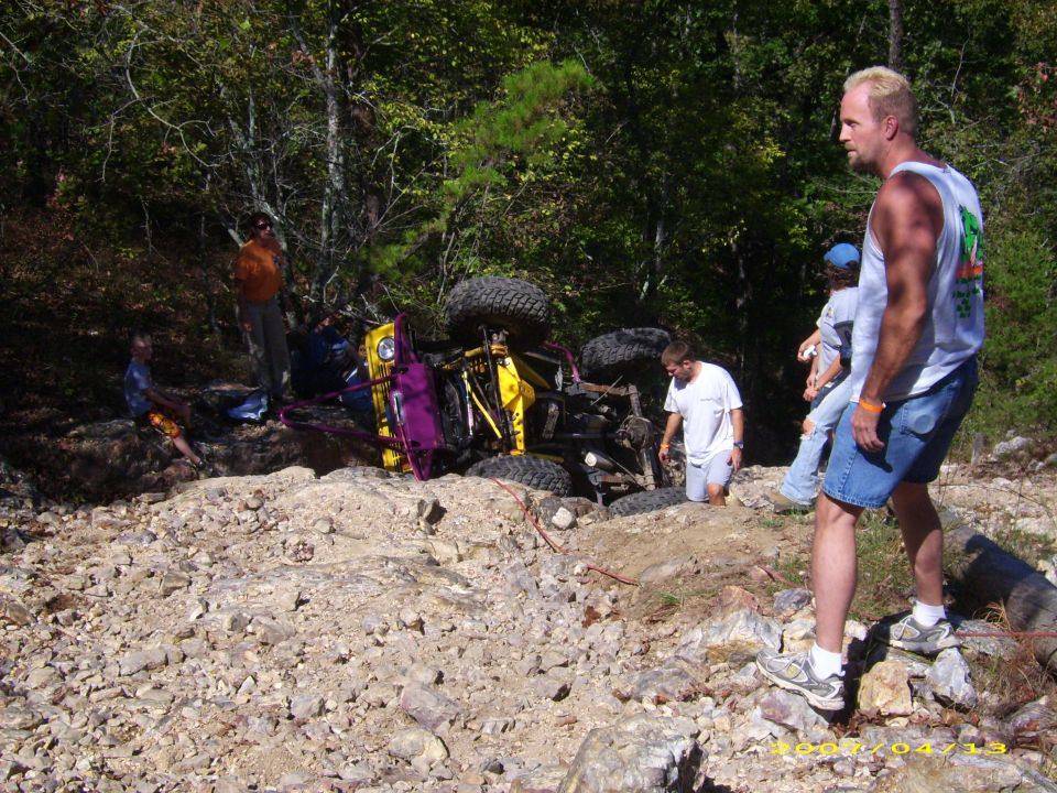 Off-road vehicle flipped on its side in a rocky, wooded area. People are standing around it