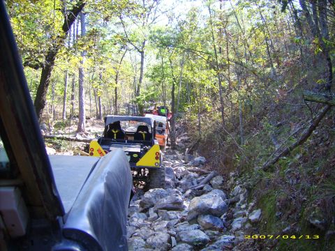 1978 Ford Bronco - Multiple off-road vehicles navigate a challenging, rocky path surrounded by dense trees.