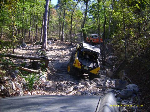 1978 Ford Bronco - Two off-road vehicles tackle a challenging rocky path through a dense forest on a sunny day.