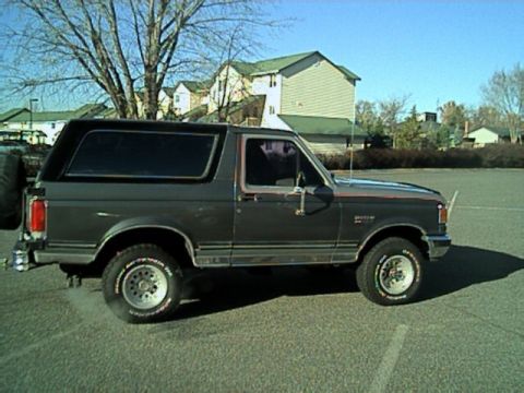 1989 Ford Bronco - A dark gray Ford Bronco is seen from the side, parked in an outdoor lot with buildings in the background.