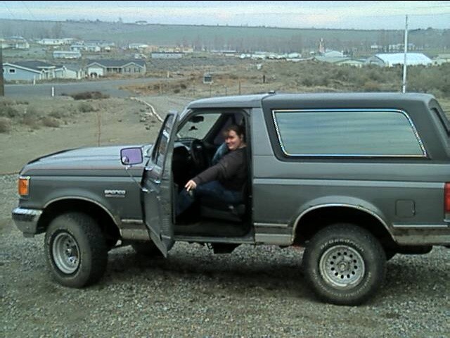 Gray two-door Ford Bronco SUV with open driver door parked on gravel, person in driver's seat