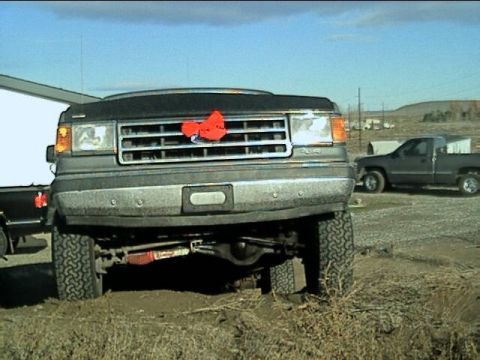 1989 Ford Bronco - Front view of a lifted Ford Bronco with a red bow on the grille; a pickup truck is visible in the background.