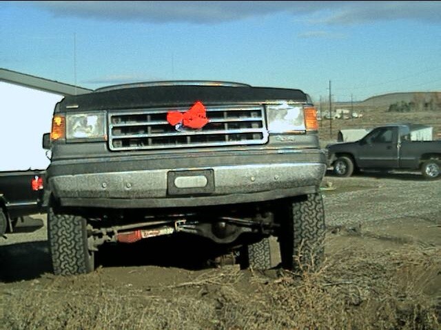 1987 Ford Bronco front end with red bow on grille. Another pickup truck is parked in the background