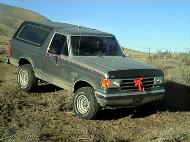 Muddy gray lifted Ford Bronco on dirt hillside, front three-quarter view with red ribbon on grille