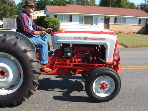 1966 Ford Ranchero - A man in overalls and a hat drives a red and white Ford 801 PowerMaster tractor down a street.