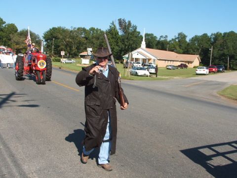 1966 Ford Ranchero - A man walks down a road during a parade, holding a camera and a rifle, with a tractor in the background.