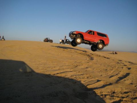 1984 Ford Bronco - A red SUV jumps over a sand dune under a clear blue sky.