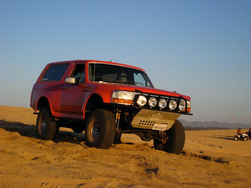 Red Ford Bronco prerunner with light bar and long-travel suspension on sand dunes at sunset
