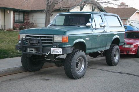 1991 Ford Bronco - This lifted 1991 Ford Bronco features a custom front bumper and winch, parked on a street with another vehicle behind.
