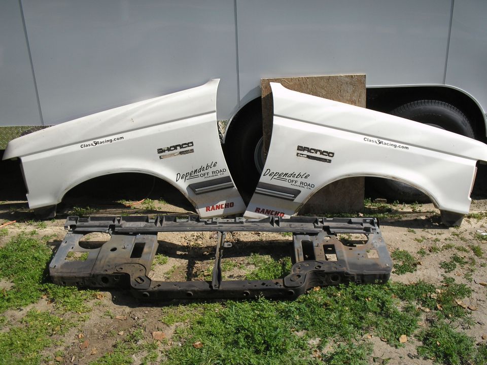 Two white Ford Bronco fenders and a black metal grille support are laid out on the ground