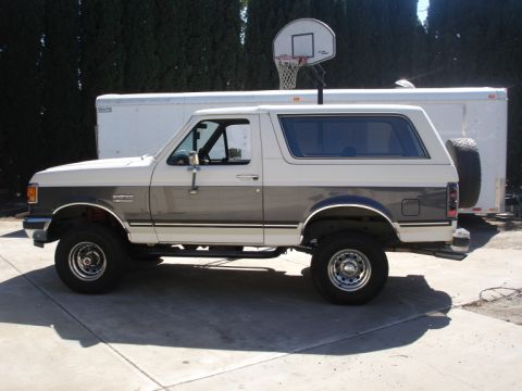 1989 Ford Bronco - This two-tone Ford Bronco is parked on a concrete driveway with a white trailer behind it.