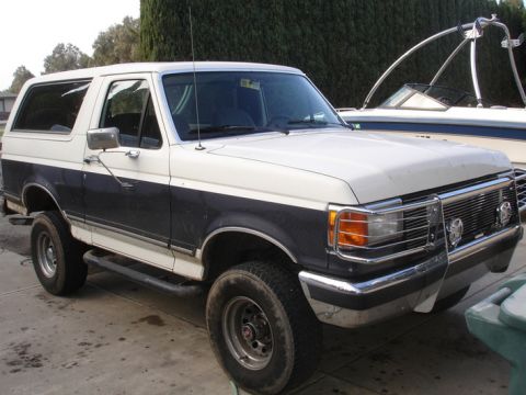 1989 Ford Bronco - This two-tone Ford Bronco is parked next to a boat, with a tree line behind it.