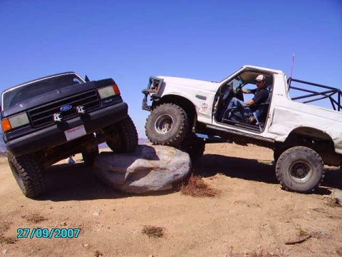 1990 Ford Bronco - Two trucks, including a 1990 Ford Bronco, tackle a challenging rocky trail under a clear blue sky.
