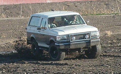1989 Ford Bronco - A lifted white-over-grey Ford Bronco with two occupants inside works through a churned-up muddy dirt field, its body flecked with mud. It h…