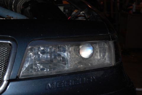 1985 Mercury Grand Marquis - Close-up of a car's headlight assembly, a projector retrofit and the 'MERCURY' badge.