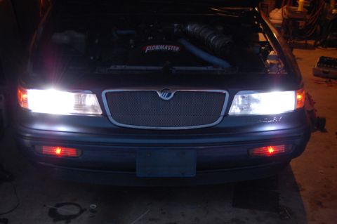 1985 Mercury Grand Marquis - Front view of a 1985 Mercury Grand Marquis, hood up, revealing engine and Flowmaster intake. Lights are illuminated.