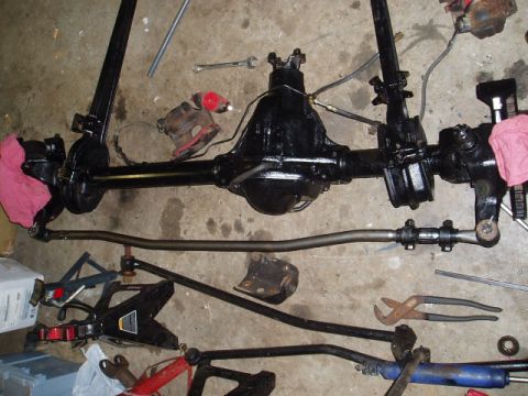 1980 Ford Bronco - Vehicle axle and steering components are disassembled and laid out on a concrete floor, with tools nearby.
