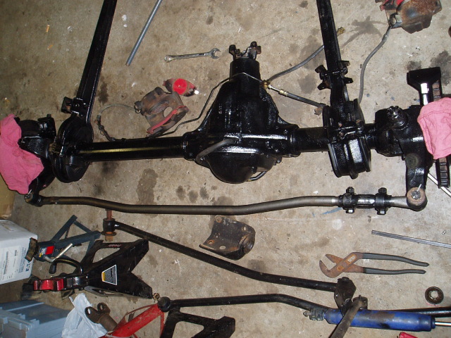 Black painted vehicle front axle assembly with steering linkage and brake components laid out on concrete floor