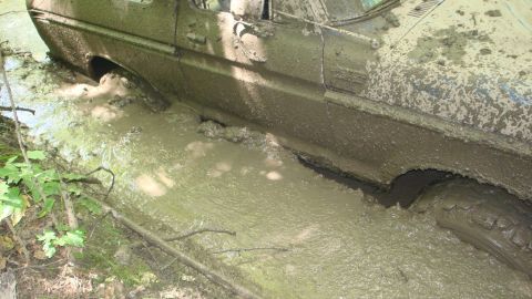 1979 Ford Bronco - A heavily mud-covered vehicle is stuck deep in a muddy ditch, with only the side and a tire visible.