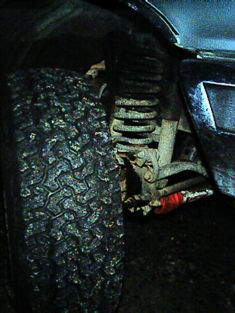 1989 Ford Bronco - Dim view up into the front wheel well past the tread of an all-terrain tire, showing the front coil spring seated on the axle perch, the co…
