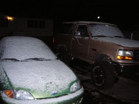 1996 Ford Bronco - Two vehicles, a 1996 Ford Bronco and a green car, sit covered in snow during the night.