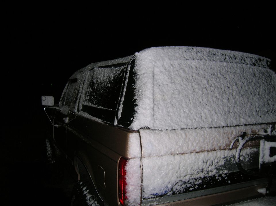 Rear view of a tan Ford Bronco covered in snow at night. Spare tire visible on the back