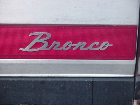 1993 Ford Bronco - Extreme close-up of a chrome script "Bronco" badge mounted on a red reflective panel set into a brushed-aluminum tailgate trim strip. A bla…