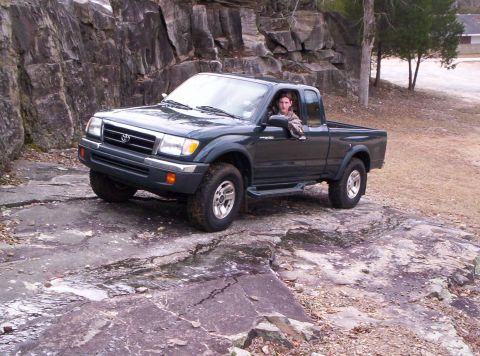 1998 Toyota Tacoma - A dark green 1998 Toyota Tacoma navigates a rocky terrain, with a driver visible through the window.