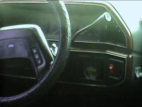 1989 Ford Bronco - Dim interior shot looking past a leather-wrapped steering wheel with a Ford-badged airbag hub at the dash, where a small round aftermarket…