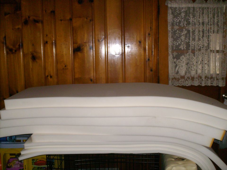 Stack of white foam padding on top of a black cage, with wood paneling and lace curtains in the background