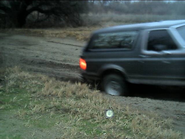 Dark full-size Bronco with brake light on, rear three-quarter, on a muddy field track at dusk