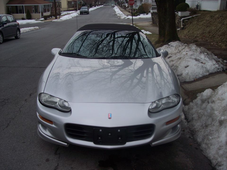 Silver convertible sports car parked on a residential street with snow on the ground. The car has a black soft top