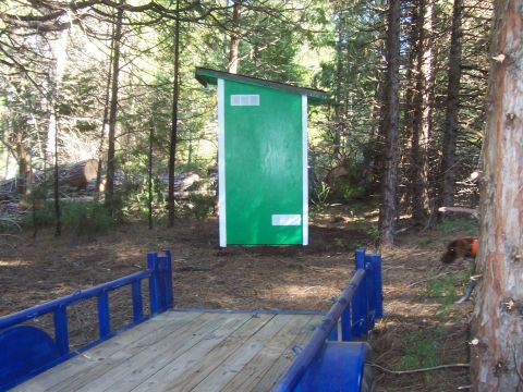 1994 Ford F-150 - A green outhouse stands in a forest clearing, with a wooden trailer parked nearby.