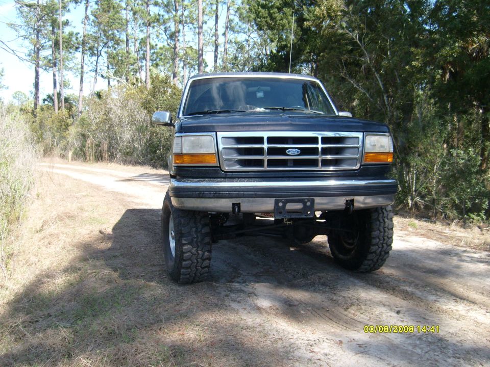 Lifted dark blue Ford Bronco on a dirt road, front view. It has a chrome grille and bumper