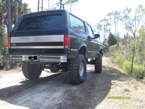 1992 Ford Bronco - Rear view of a black 1992 Ford Bronco with a silver stripe and lifted suspension on a dirt path.
