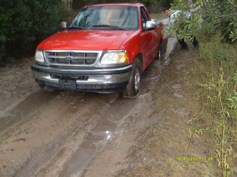 1992 Ford Bronco - A red Ford F-150 is stuck in a muddy track, with a person attempting to push it free.
