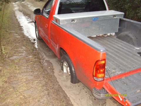 1992 Ford Bronco - This red pickup truck appears to be stuck in the mud on a dirt road, with its rear wheel submerged.