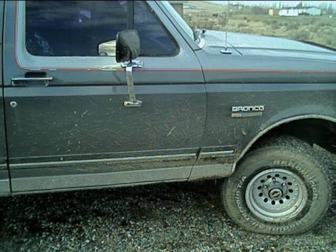 1989 Ford Bronco - Close side view of a gray Ford Bronco's driver door and front fender caked in dried mud splatter, with the 'BRONCO' fender badge, a red pin…