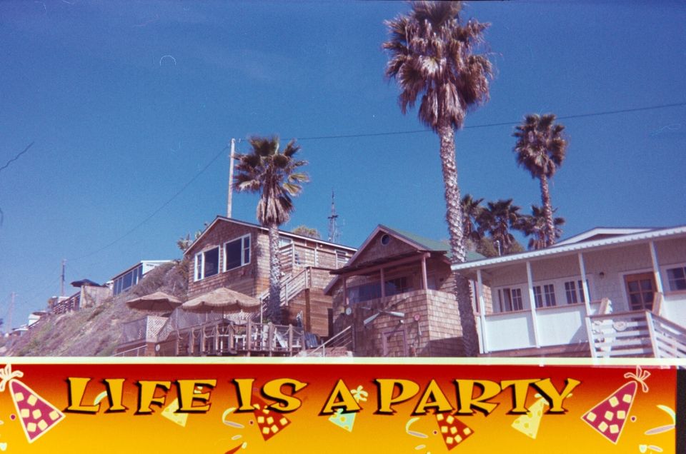 Beachside buildings with palm trees under a clear blue sky. A banner reads "LIFE IS A PARTY"
