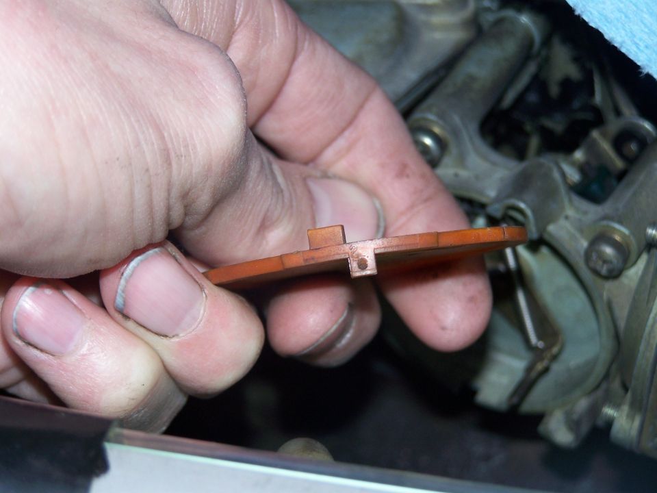 Hand holding a small, orange, rectangular carburetor choke component with a small hole and a tab