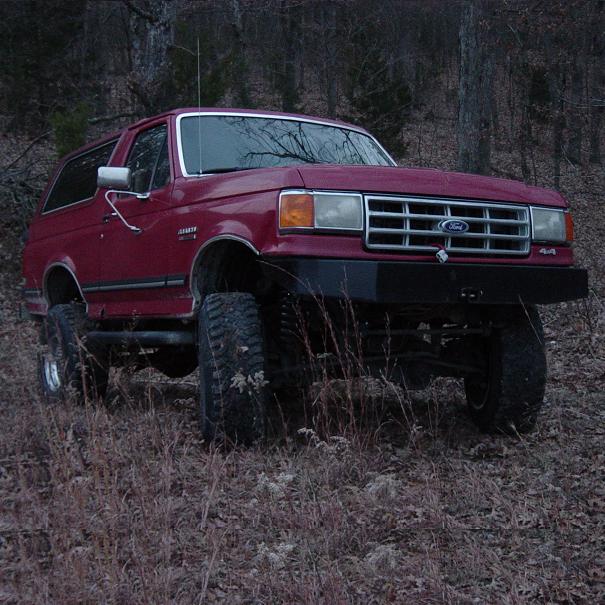 Red lifted Ford Bronco front three-quarter in dim wooded field, chrome grille and 4x4 badge