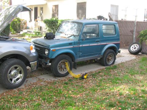 1987 Suzuki Samurai - A teal Suzuki Samurai is parked next to a gray pickup truck, with a flat tire being inflated by a portable air compressor.