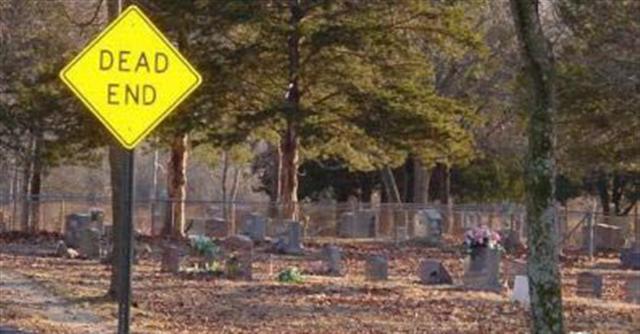 Yellow diamond dead end sign in front of a cemetery with gravestones and trees