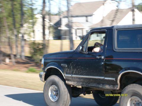1992 Ford Bronco - A black Ford Bronco is seen driving on a road with houses and trees in the background. The vehicle has mud splatters on its side.