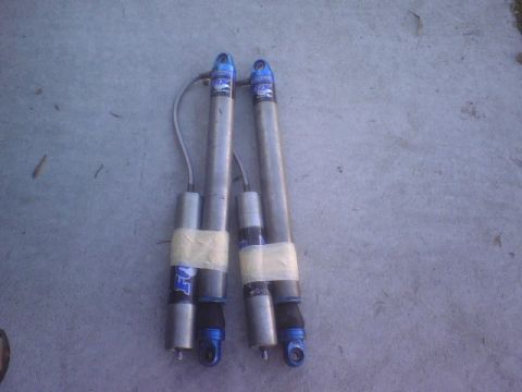 1984 Ford Bronco - Two Fox shock absorbers are laid out on a concrete surface, showing their blue and silver components.