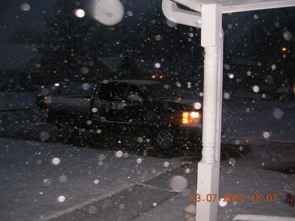Dark pickup truck with headlights on parked in a snowy driveway during a night snowstorm