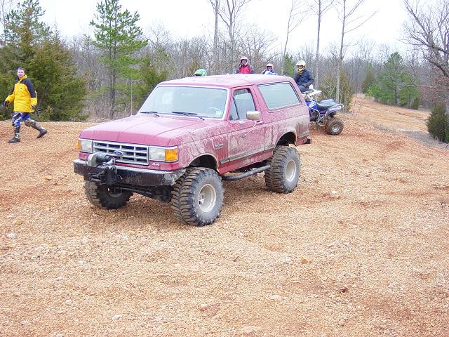Mud-splattered red lifted Ford Bronco parked on a dirt hill with ATV riders nearby