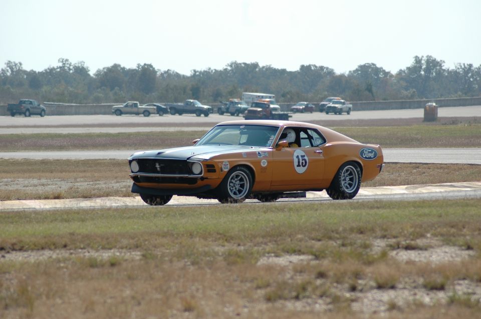 Orange No. 15 Mustang race car on track with paddock trucks in the background