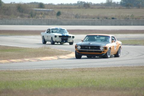 1970 Ford Mustang - Two Ford Mustangs compete on a race track, with the orange and black model in the foreground.