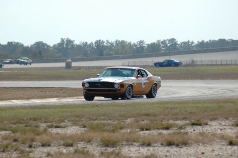 1970 Ford Mustang - A classic orange Mustang competes on a race track with other vehicles in the background.